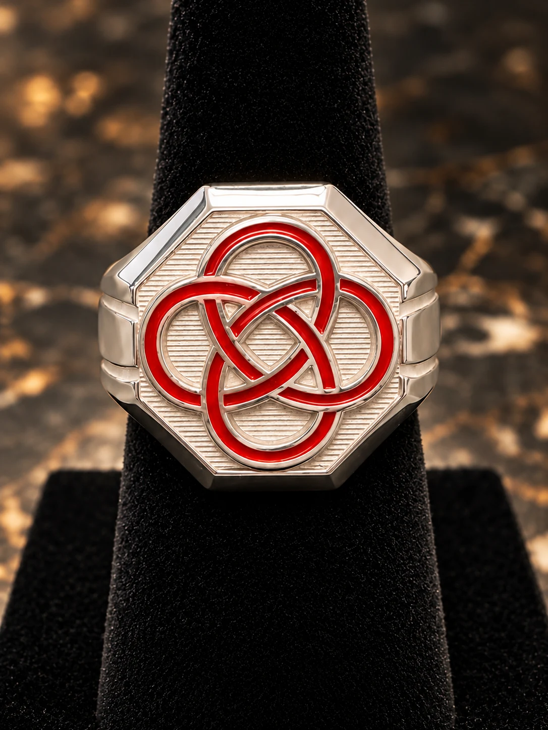 Silver octagonal signet ring with an interwoven red enamel knot and a horizontally textured face, on a black velvet stand